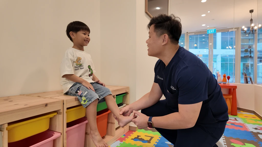 A podiatrist checking the bare foot of a smiling young child seated in a clinic play area, the assessment that guides podiatrist recommended shoes for kids.
