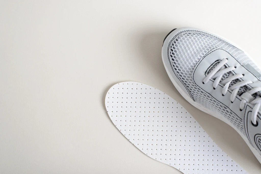 A grey mesh running shoe beside the flat perforated insole taken out of it on a pale background, the mass-made liner side of the orthotics vs insoles question.