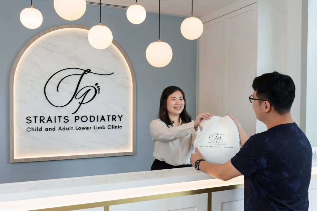 Straits Podiatry receptionist handing a branded tote bag to a patient across the marble reception counter, beneath the arched Straits Podiatry logo wall.