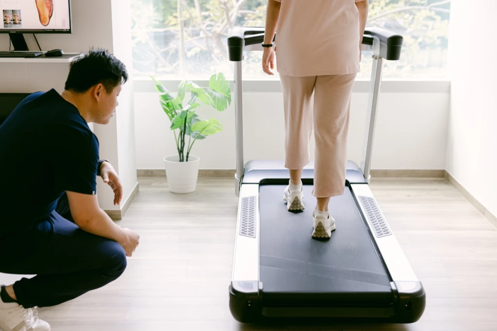 Podiatrist observing a patient walk on a treadmill during gait analysis, with foot-scan results on the screen behind