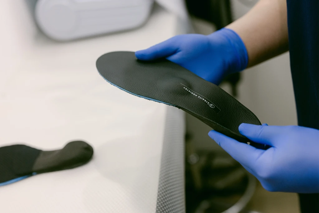 A gloved podiatrist holding a black custom insole branded Straits Podiatry, showing its durable top cover