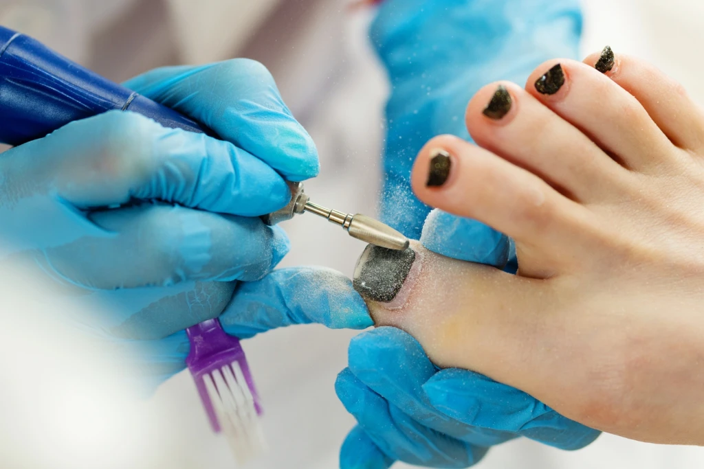 A pedicurist in blue gloves using an electric nail drill on a polished toenail during a salon pedicure