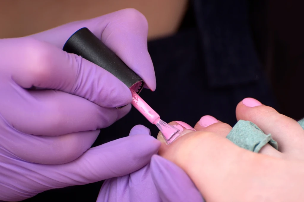 A pedicurist in gloves applying pink nail polish over a toenail during a pedicure