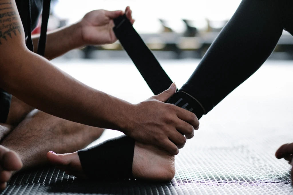 Hands wrapping a black elastic support strap around a person's bare ankle and foot on a gym mat, external ankle support during rehabilitation