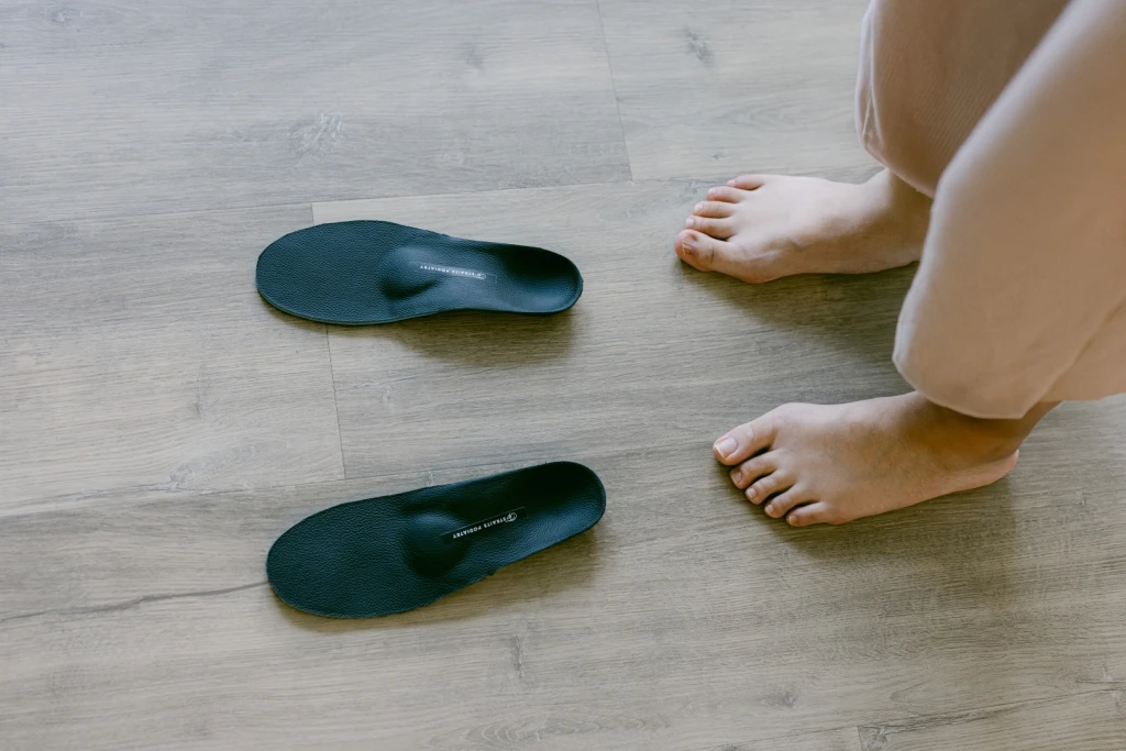 A pair of customised orthotic insoles on a wooden floor, to be fitted for a person who has a bunion on each foot, seated barefoot beside them